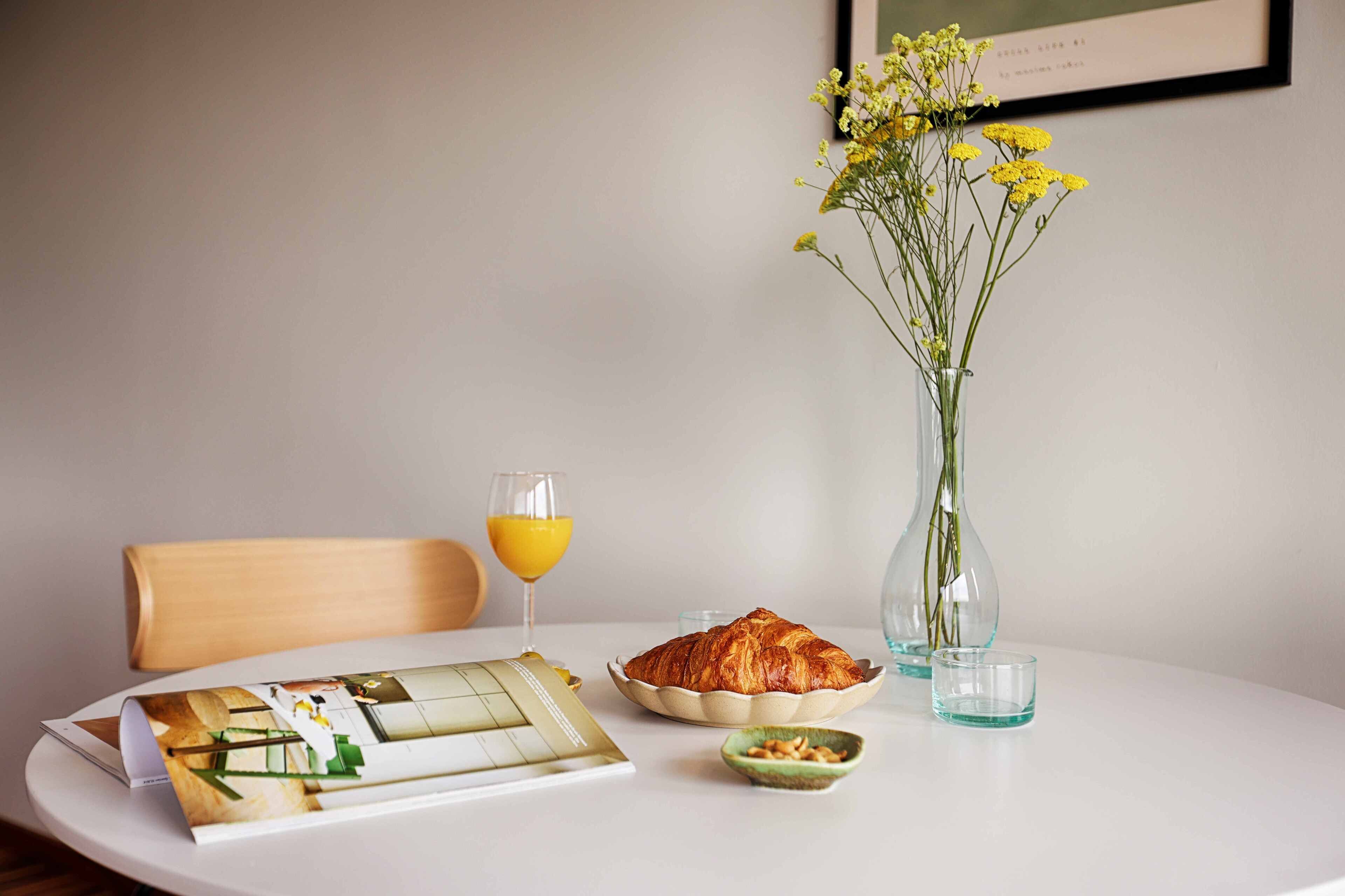 A breakfast setting with a croissant, a glass of orange juice, a vase with yellow flowers, and a menu on a white table.