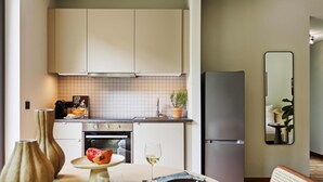 A modern kitchen with white cabinets, a tiled backsplash, a built-in oven, and a refrigerator. There is a dining table with a vase, a plate, and a glass of wine.