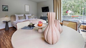 A modern hotel room with a bed, a dining table with a vase, and a view of greenery outside.