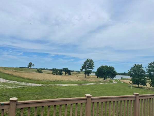 Un campo cubierto de hierba con una valla de madera en primer plano, una masa de agua al fondo y un cielo despejado.