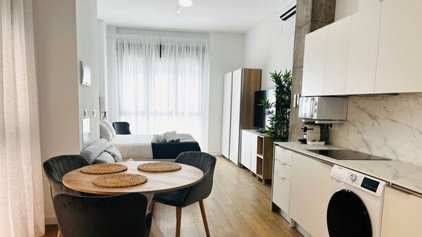 A modern kitchen with a built-in washing machine, a dining area with a round table and chairs, and a bedroom with a bed and curtains. - A (VA070.2.4) TH Mediterranean Suites 4 (Valencia / València)