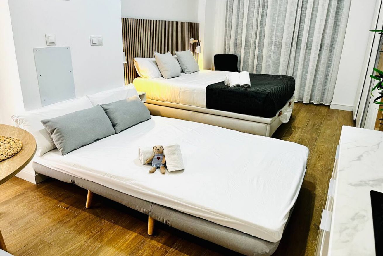 A hotel room with two beds, a wooden floor, a white wall, a window with curtains, and a small table with a teddy bear.