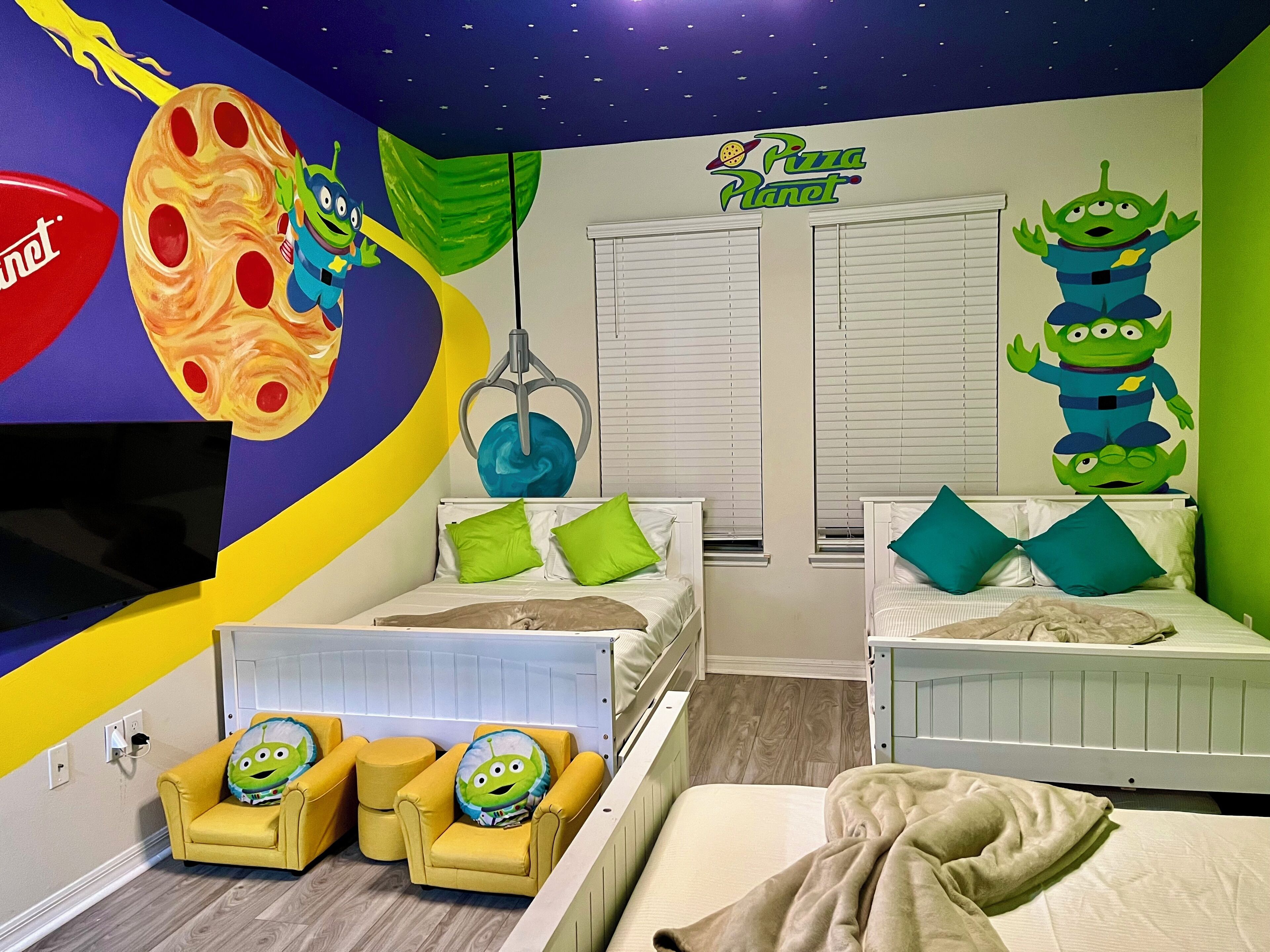Een kinderkamer met een ruimtethema muurschildering, twee bedden, een televisie en een bank met een groen alien kussen.