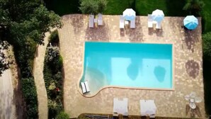 Vue aérienne d’une piscine dotée d’une terrasse en pierre, entourée de verdure et aménagée avec des chaises longues et des parasols.