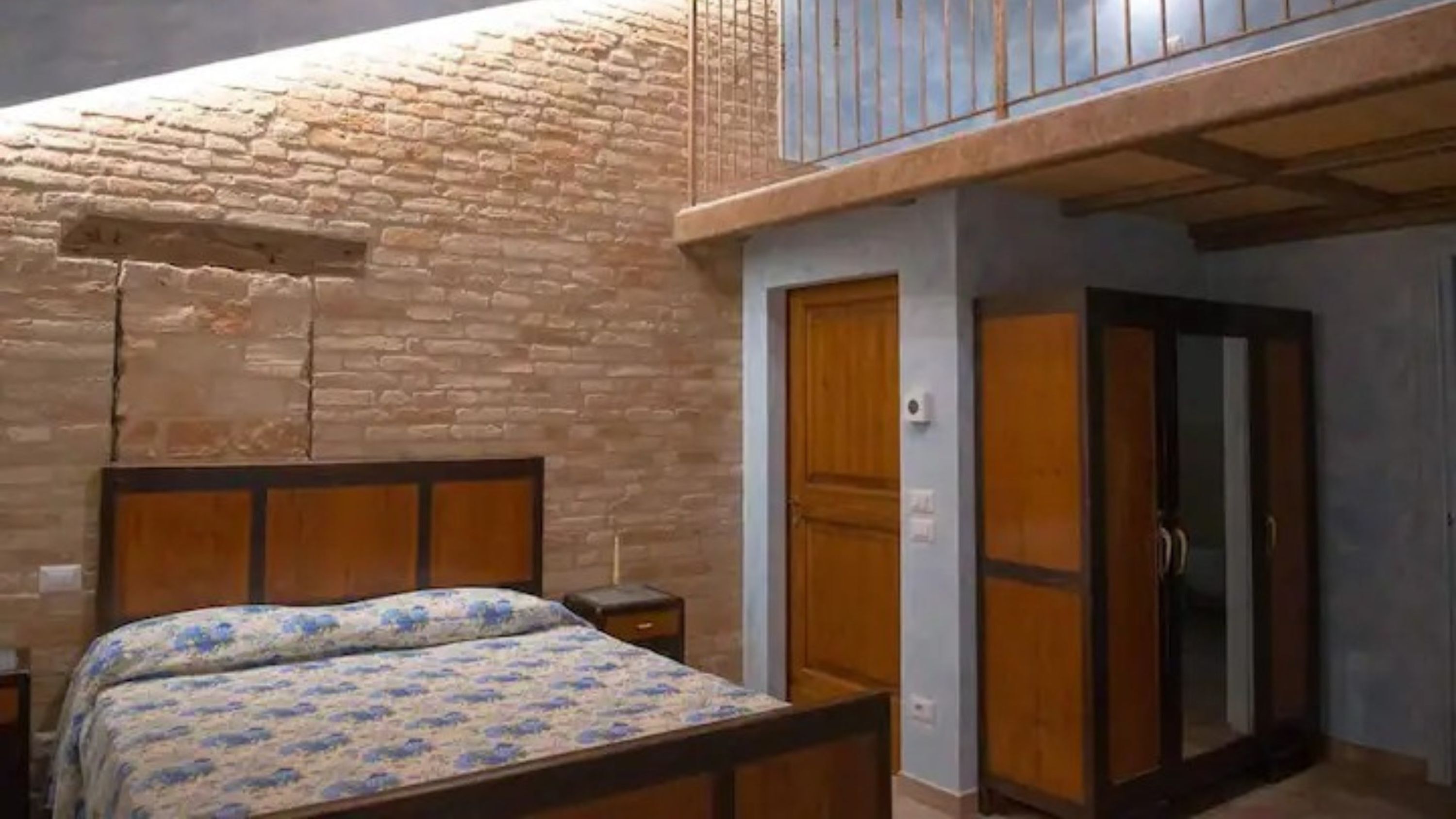 Quarto com cama, cabeceira de madeira, uma pequena mesa de cabeceira, um guarda-roupa e uma varanda com corrimão.