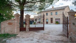 Une entrée fermée par un portail, avec un mur en pierre et un arbre, qui mène à un bâtiment doté d’un toit en tuiles et de nombreuses fenêtres.