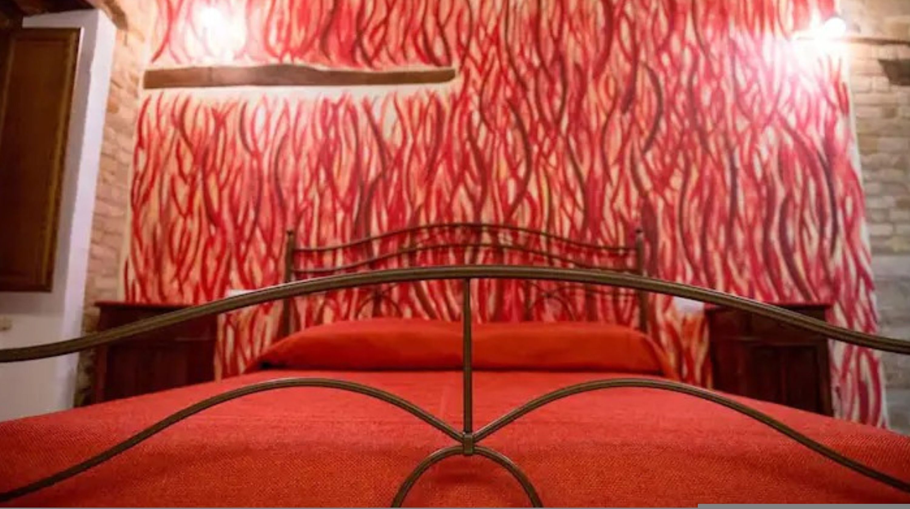 A bed with a metal frame and red bedding in a room with a red patterned wall and a wooden shelf above it.