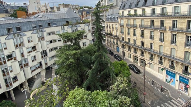 Un immeuble à plusieurs étages doté de balcons, une rue au premier plan et un mélange de bâtiments résidentiels et commerciaux en arrière-plan.