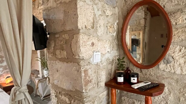 A room with a stone wall, a wooden console table with a mirror and candles, a TV mounted on the wall, and a curtain tied to a chair. - Eskikent Alaçatı (Cesme)