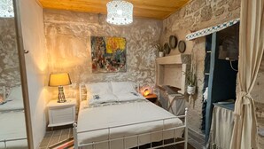 A bedroom with a stone wall, a bed with white bedding, a bedside lamp, a colorful rug, and a hanging plant. - Eskikent Alaçatı (Cesme)
