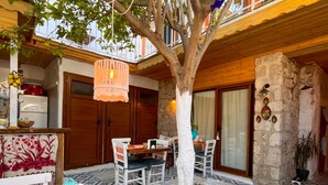 A cozy outdoor seating area with a wooden table and chairs, surrounded by a stone wall and a tree. - Eskikent Alaçatı (Cesme)