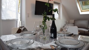 A dining table set with plates, glasses, and cutlery, a vase with a rose, and a bottle of wine, with a TV and a couch in the background. - "Le Rooftop de la fontaine", in a quiet, green setting (Luisant)