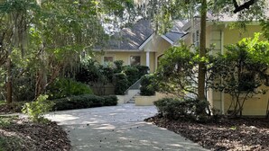 A residential house with a paved driveway leading to a front door, surrounded by lush greenery and trees.