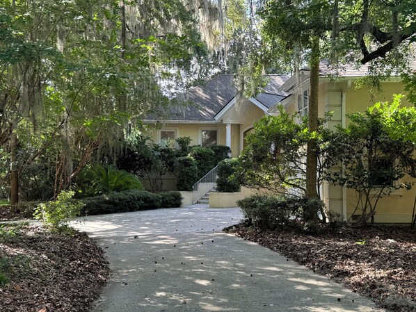 A residential house with a paved driveway leading to a front door, surrounded by lush greenery and trees.