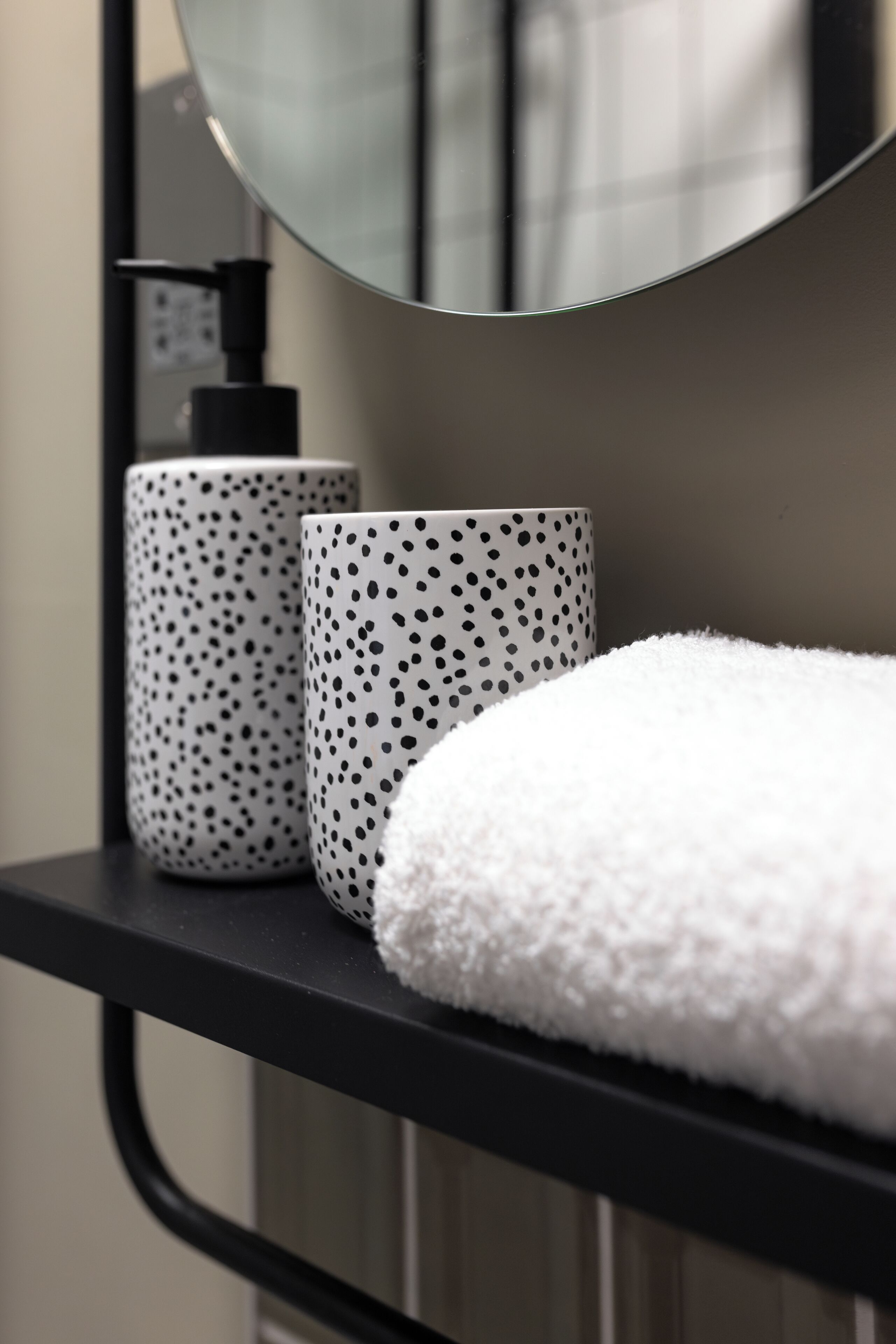A bathroom with a black shelf holding two white mugs with black dots, a white fluffy towel, and a mirror above.