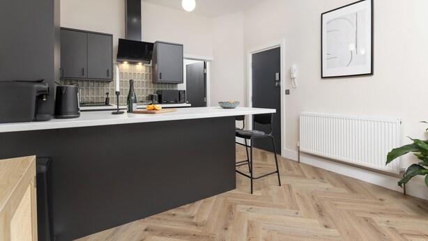 A modern kitchen with a central island, black cabinets, and a white countertop. There is a black chair and a potted plant in the corner. - Broughton Place (Liverpool)