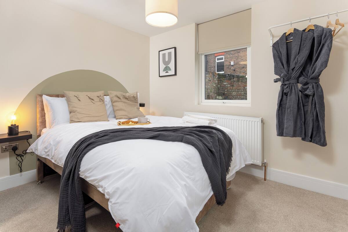 A neatly made bed with a white duvet, a grey throw blanket, and a beige pillow. There is a framed picture on the wall, a radiator, and a window with a view of a brick building.
