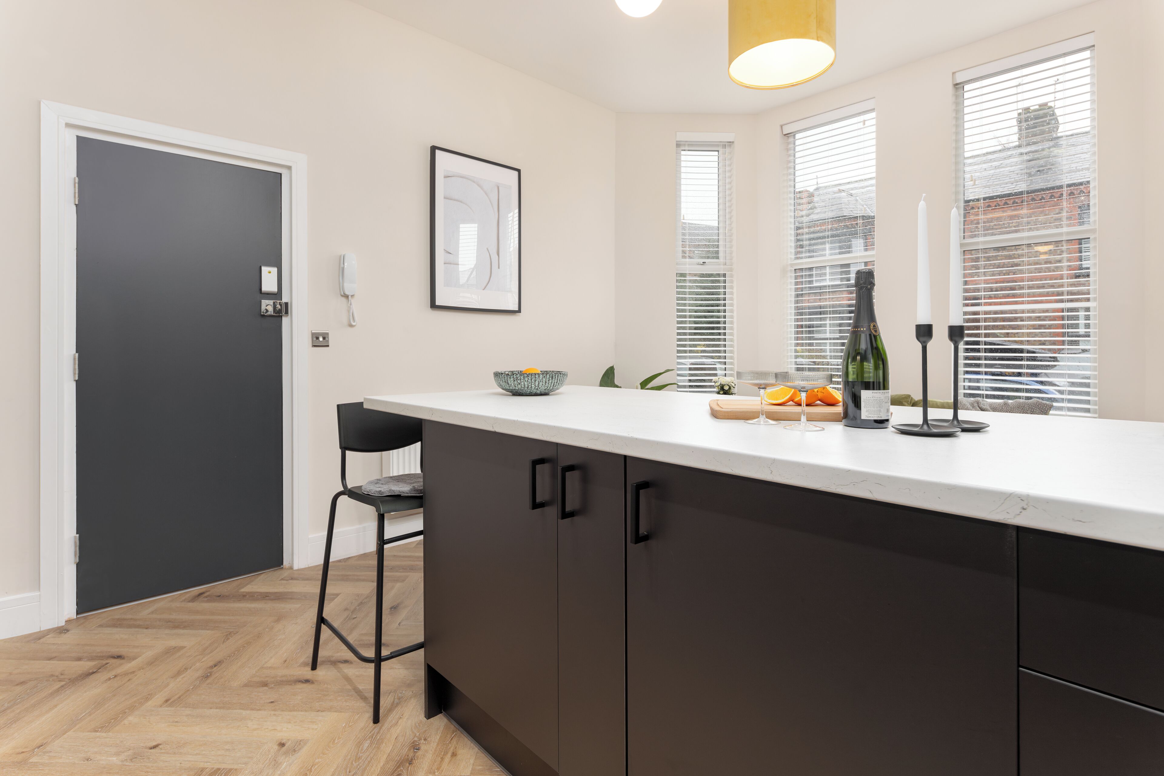 A modern kitchen with a white countertop, dark cabinets, a black chair, a framed picture, and a window with blinds.