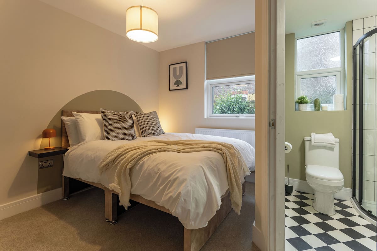 A bedroom with a bed, bedside table, lamp, and a window with a view of greenery.