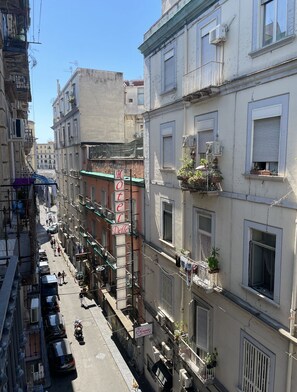 Una calle estrecha bordeada de edificios de varios pisos, algunos con balcones y unidades de aire acondicionado. Se ve un cartel de un hotel, y hay coches aparcados y un motociclista en la calle.