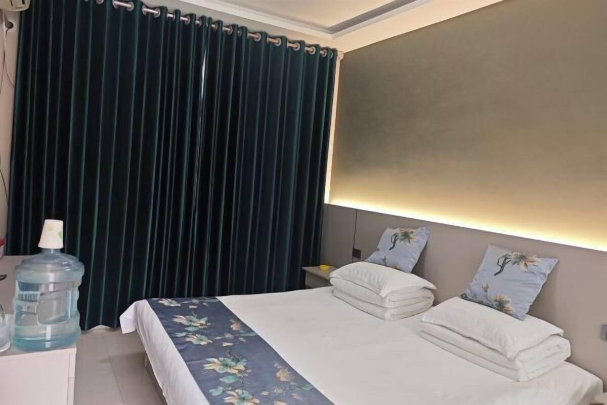 Hotellihuone, jossa on sänky, yöpöytä, vesiautomaatti ja verho.