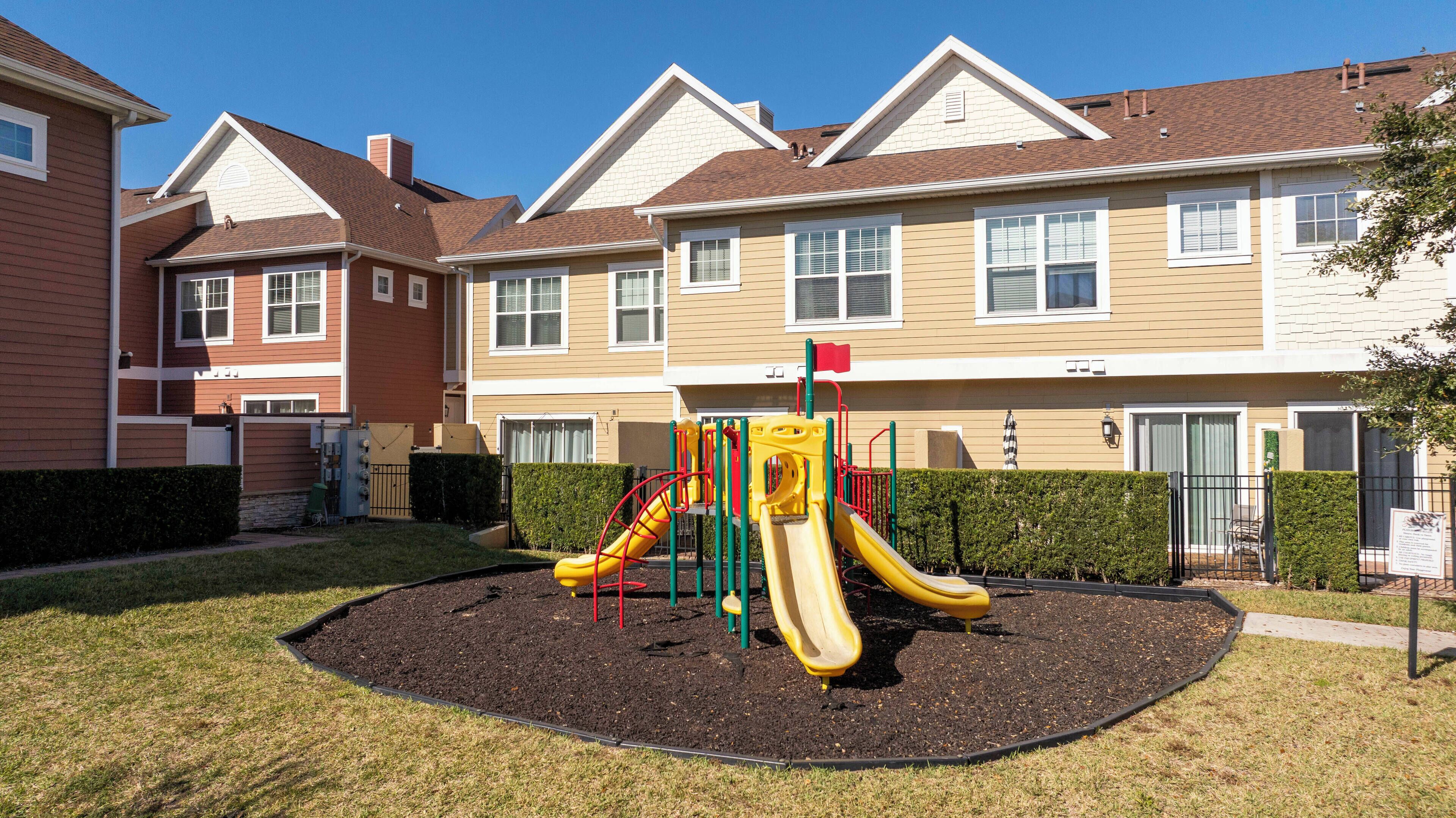 A playground with a yellow slide and a red climbing structure, surrounded by a well-maintained lawn and a fence.