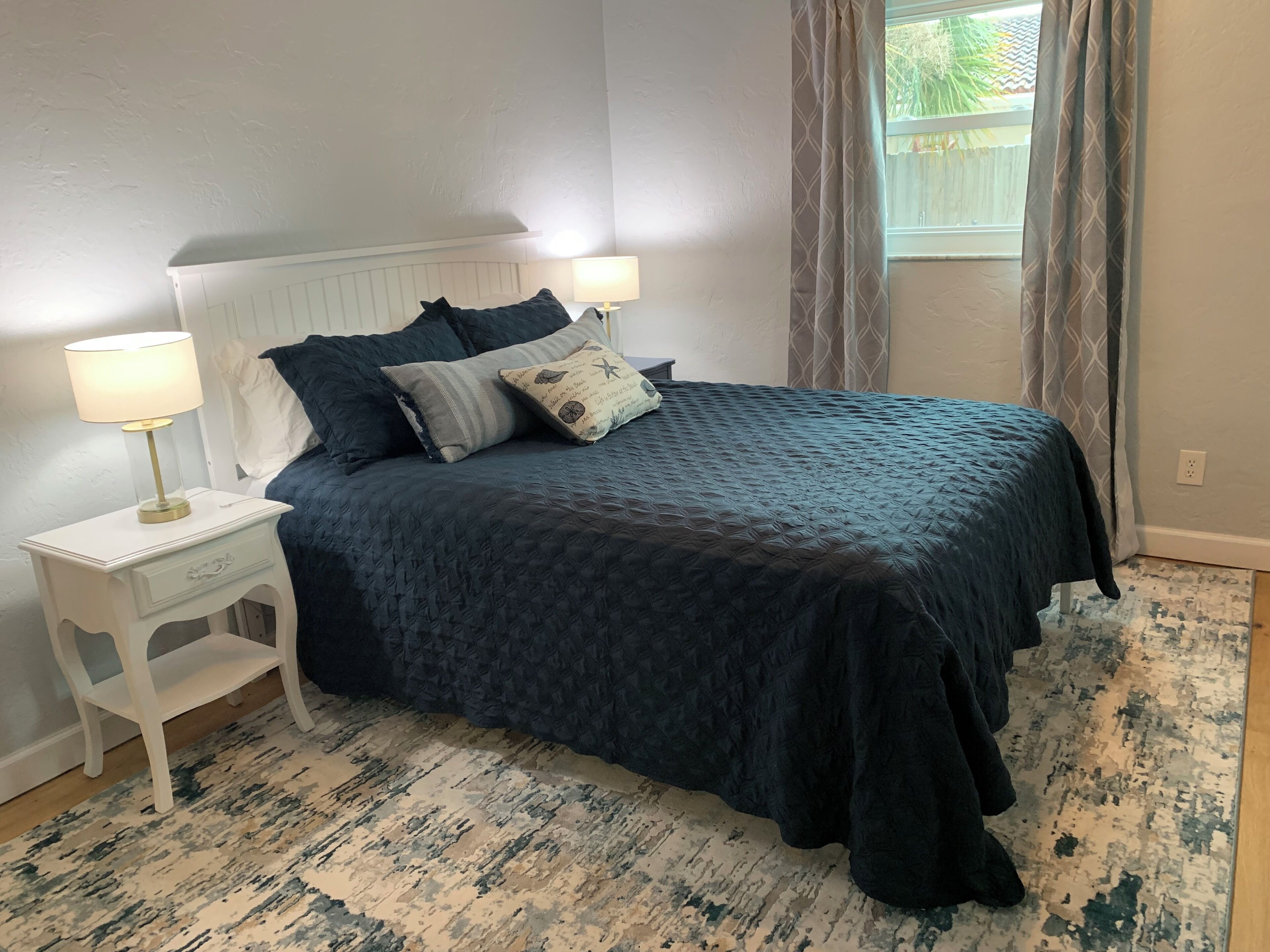 A bedroom with a dark blue bedspread, two bedside lamps, a white nightstand, and a patterned rug.