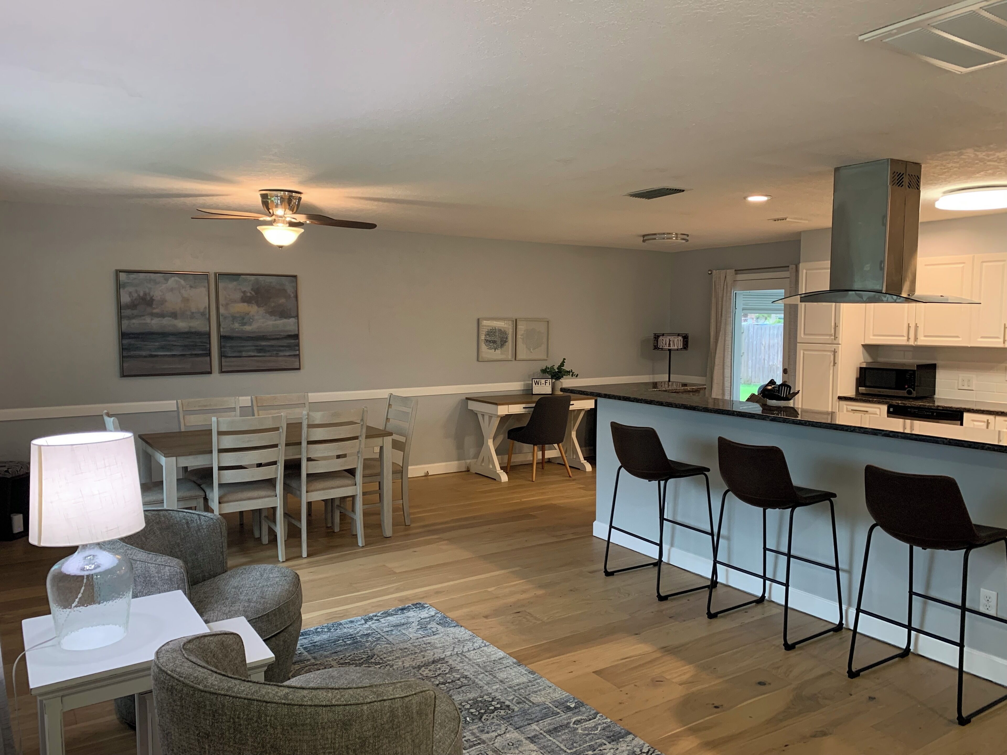 A modern kitchen with white cabinets, a bar area with stools, a dining table with chairs, and a ceiling fan.