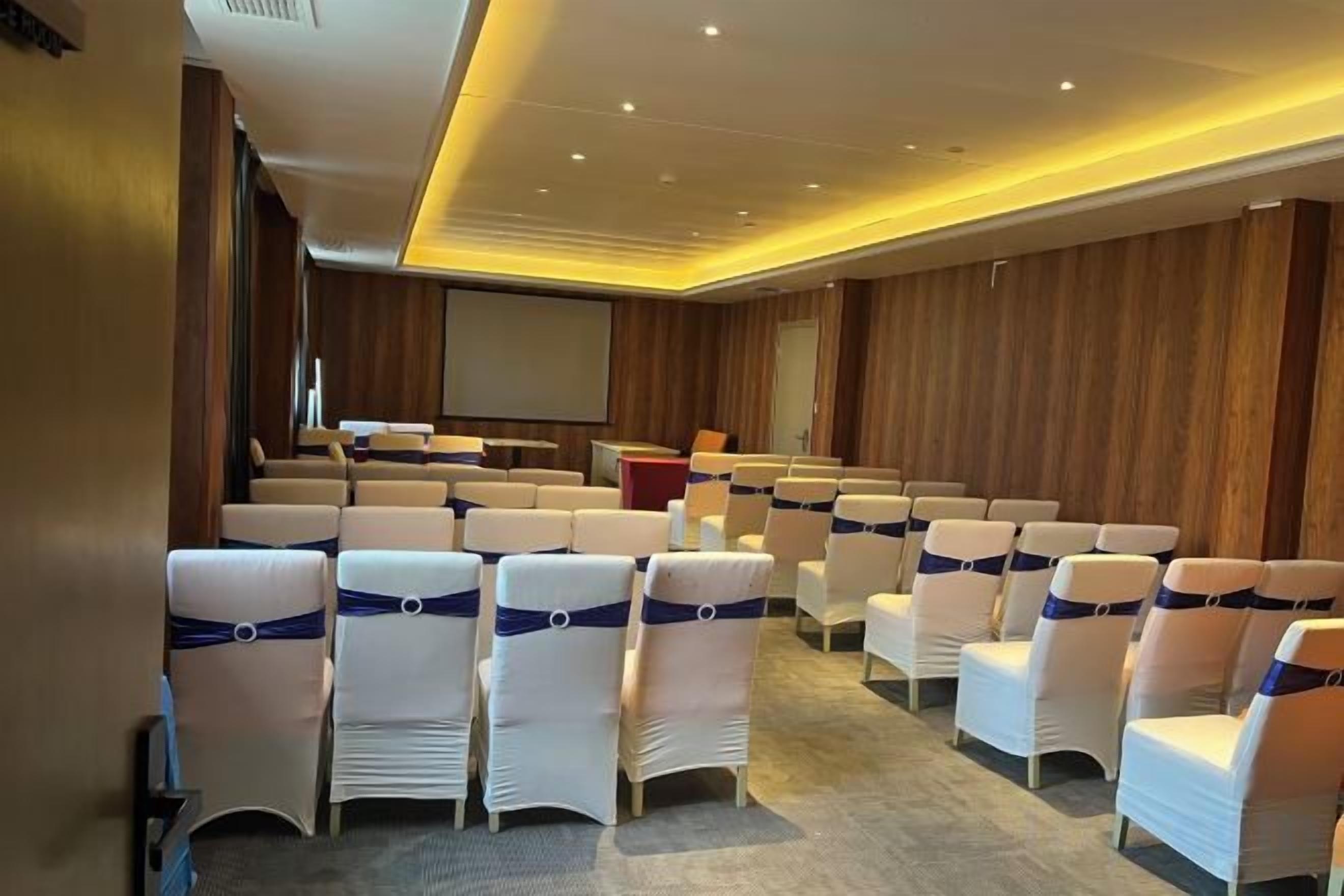 A conference room with rows of white chairs, a podium, and a large screen.