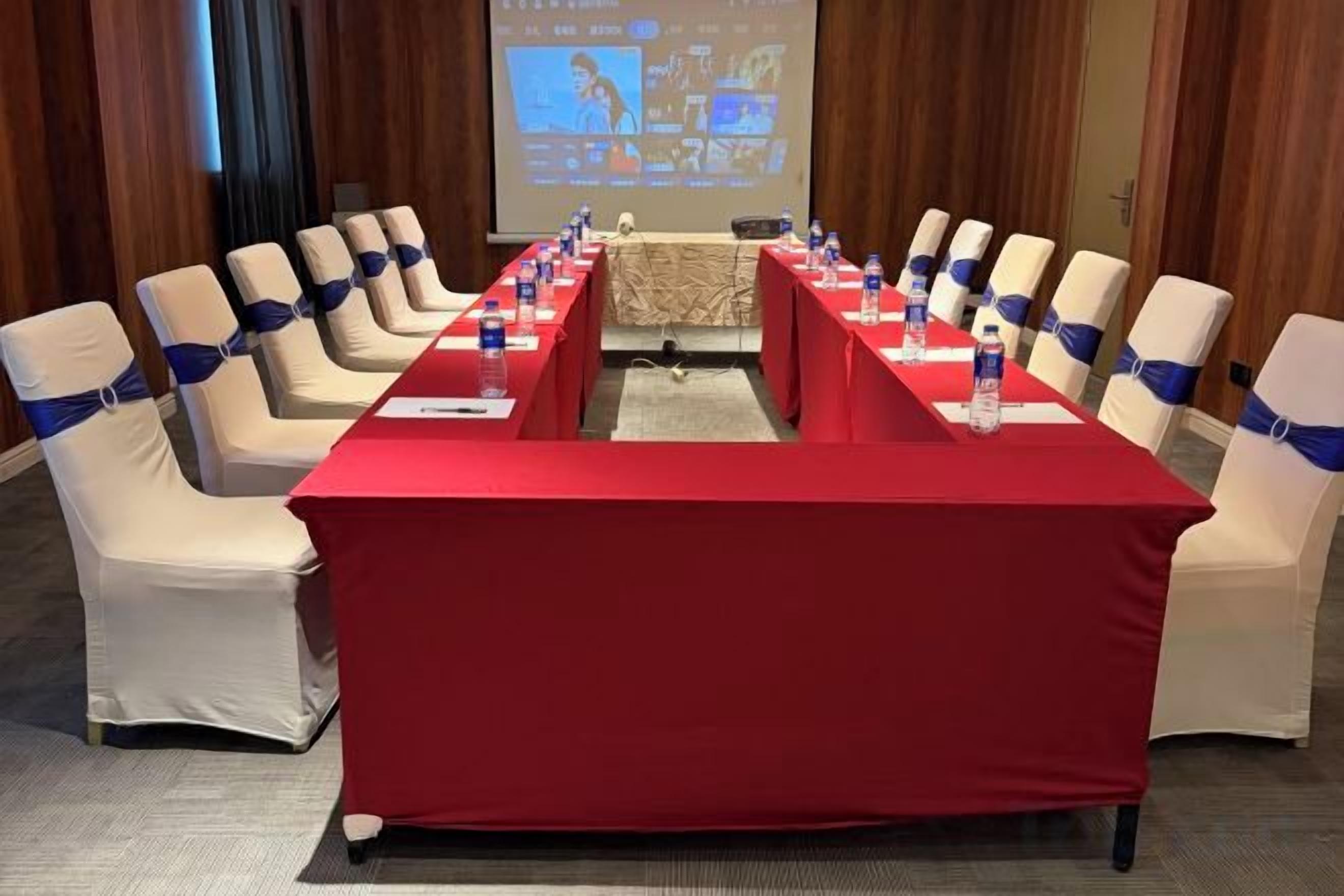 Un salón de conferencias con una larga mesa roja, sillas color beige con cintas azules y una pantalla de proyección al fondo.