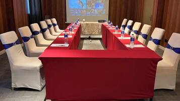 Un salón de conferencias con una larga mesa roja, sillas color beige con cintas azules y una pantalla de proyección al fondo.