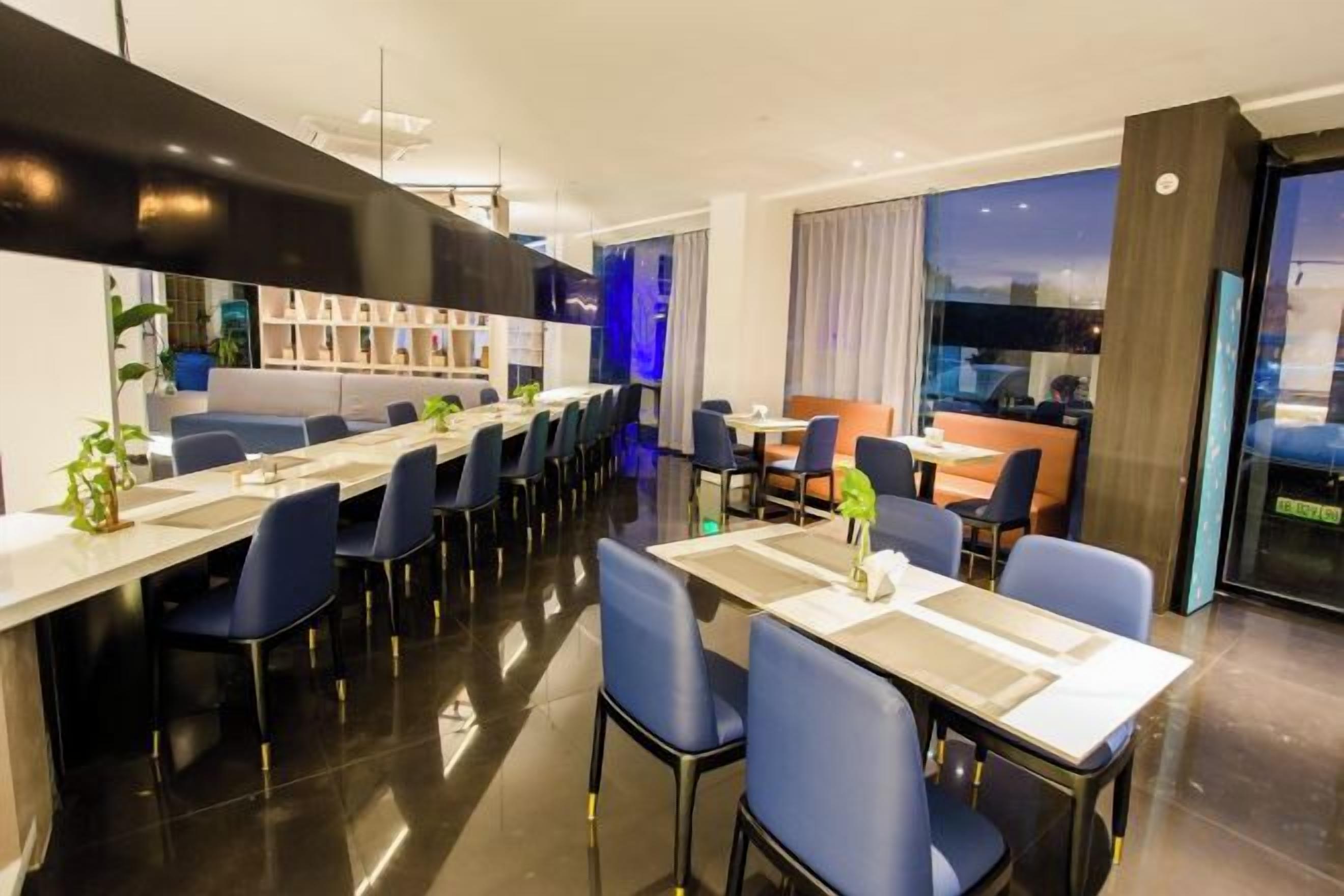 Un comedor moderno de hotel con mesas y sillas largas, una zona de bar y vistas a la ciudad.