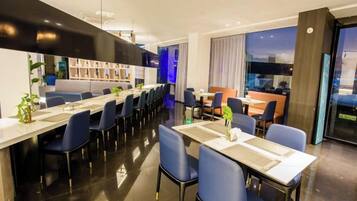 Un comedor moderno de hotel con mesas y sillas largas, una zona de bar y vistas a la ciudad.
