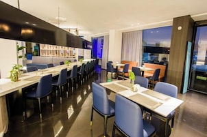 Un comedor moderno de hotel con mesas y sillas largas, una zona de bar y vistas a la ciudad.