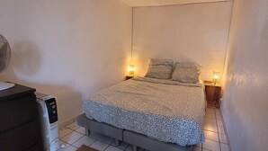 Une petite chambre à coucher, bien rangée, avec un lit, deux lampes de chevet, un radiateur et un ventilateur.