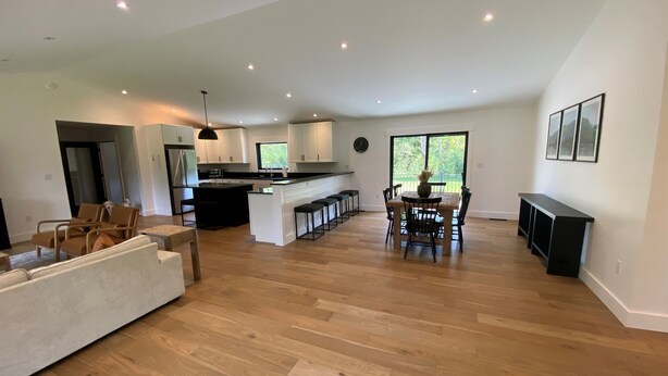 A spacious living area with a wooden floor, a dining table with chairs, a kitchen with white cabinets, and a large window providing a view of the outdoors. - Escape to The Clyde’s View Hideaway in Manchester, Vt. New Home  3 Bed 2 Bath (Manchester Center)