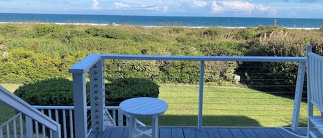 A wooden deck with a white railing, a small table, and a chair. The deck overlooks a grassy area with dense vegetation and a view of the ocean.