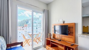 Una habitación con cama, un televisor sobre un mueble de madera, una puerta corrediza de vidrio a un balcón y una mesita con un florero.