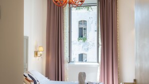A hotel room with a bed, a chandelier, a window with curtains, and a wall-mounted lamp.