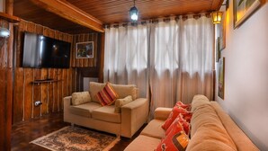 A cozy living room with a wooden ceiling, a flat-screen TV mounted on the wall, a beige sofa with patterned cushions, a red throw blanket, and a patterned rug on the wooden floor. - Ap in Capivari - Tirol31 By Temporada Férias (Campos do Jordão)