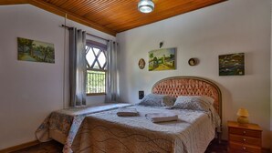 A bedroom with a wooden ceiling, a bed with a patterned bedspread, a nightstand with a lamp, and artwork on the walls. - Ap in Capivari - Tirol31 By Temporada Férias (Campos do Jordão)