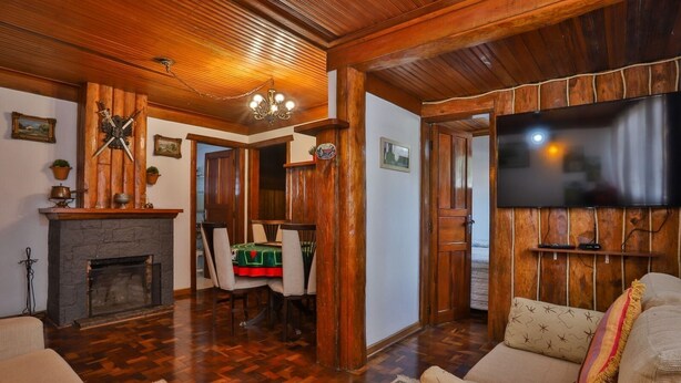 A cozy living room with a fireplace, wooden walls, a TV mounted on the wall, and a dining area with a table and chairs. - Ap in Capivari - Tirol31 By Temporada Férias (Campos do Jordão)