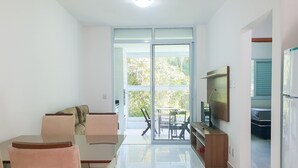 A modern living room with a sofa, dining table, and chairs. There is a TV on the wall and a sliding glass door leading to a balcony.