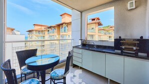 En balkon med et rundt bord og stole, et tekøkken med grill, og udsigt til nabobygninger.