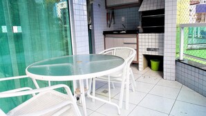 Un pequeño patio con una mesa redonda de vidrio y sillas de mimbre blanco, un salpicadero de azulejos y vista a través de una cerca de malla verde.