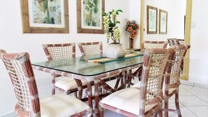 Un comedor con una mesa de vidrio rodeada de sillas de mimbre con cojines blancos. En las paredes, hay cuadros enmarcados con motivos botánicos y un espejo con marco tejido.