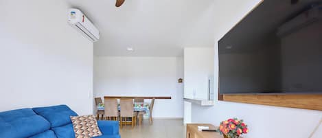 Una sala de estar con un sofá azul, una mesa de comedor con sillas, un televisor de pantalla plana montado en la pared y una pequeña mesita de madera con un jarrón de flores.