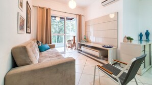 A living room with a sofa, a dining table, and a chair. There is a window with curtains, a ceiling light, and an air conditioning unit. - Apartment at Big Beach TF305 By Temporada Férias (Ubatuba)