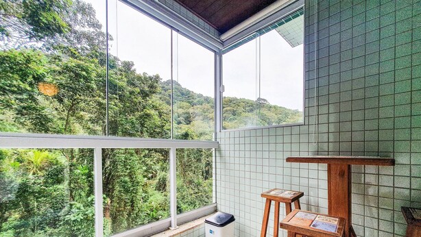 A balcony with a view of lush greenery, a tiled wall, and a small wooden table with stools. - Apartment at Big Beach TF305 By Temporada Férias (Ubatuba)