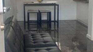 Een woonkamer met een zwarte bank, een kleine tafel met twee stoelen, een raam met zonwering, en een keuken met kasten.